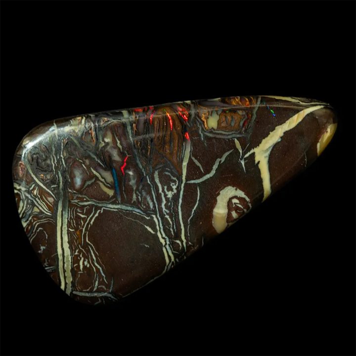 Opale naturelle australienne des mines Koroit. Forme triangulaire de 14,4 carats à matrice sombre brun sombre homogène bariolée de dessins de couleurs claires crème et gris-bleu ; certaines plages longilignes s’irisent de rouge sang, bleu et vert brillants